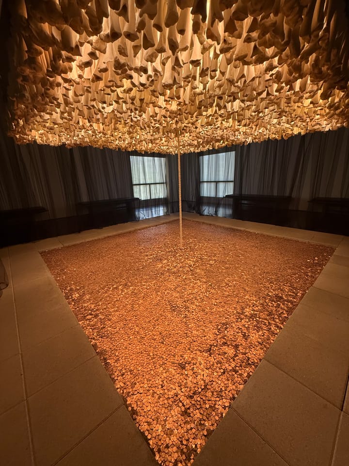 Installation view: a vast carpet of copper coins on a gallery floor, linked by a thin column of wafers to a canopy of hanging cattle bones.